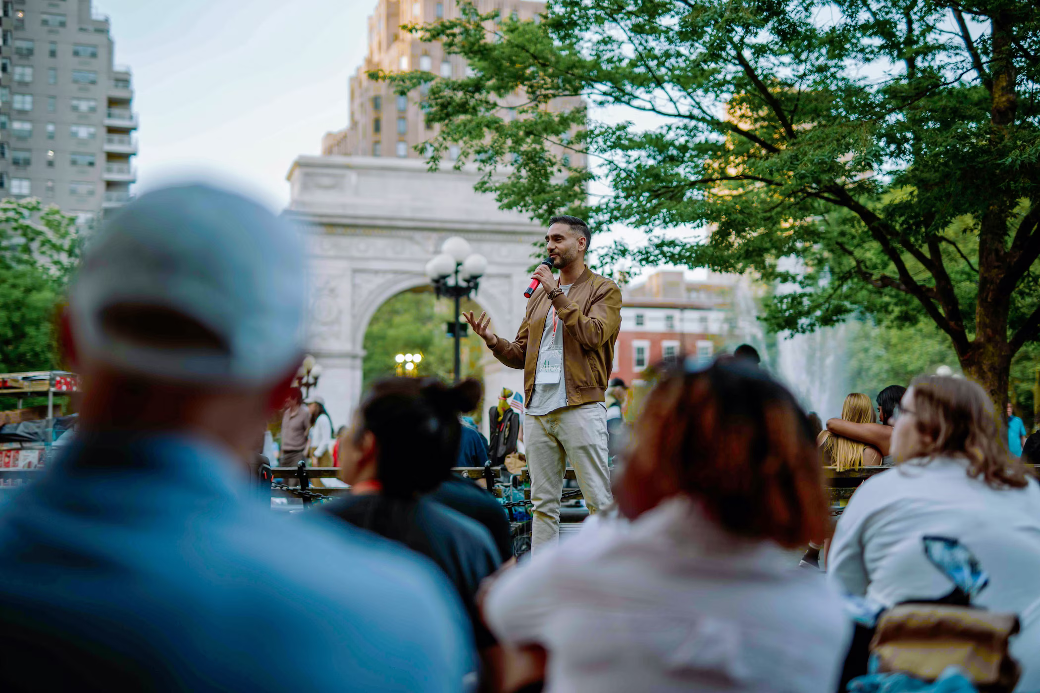Man speaking into a microphone in a park with a crowd seated in front of him and an arch monument in the background.