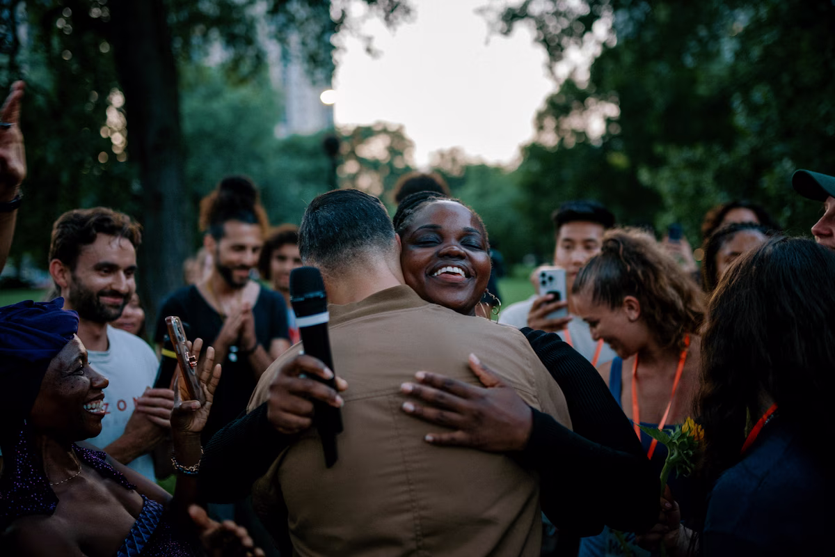 A joyful woman hugging a man holding a microphone during an outdoor gathering with people smiling and clapping around them.