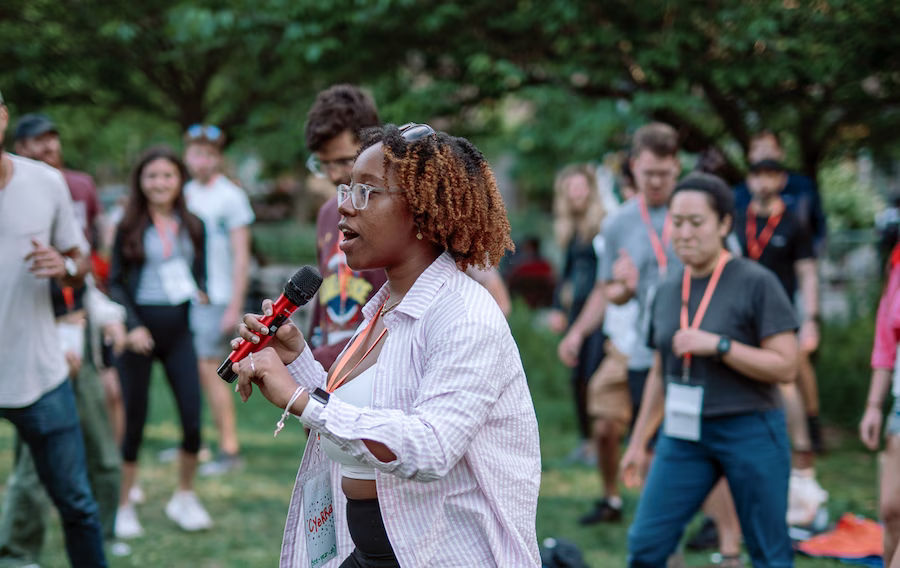 Woman speaking into a microphone outdoors in front of a group of people at an event.