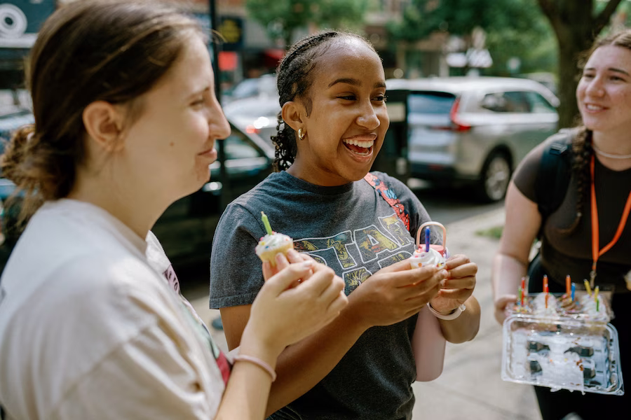 Three young women outdoors smiling and holding cupcakes with lit candles, one woman holds a plastic container with more cupcakes.
