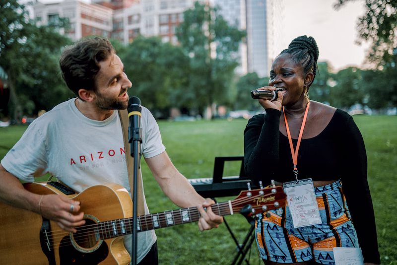 A man playing acoustic guitar and a woman singing outdoors in a park with buildings and trees in the background.