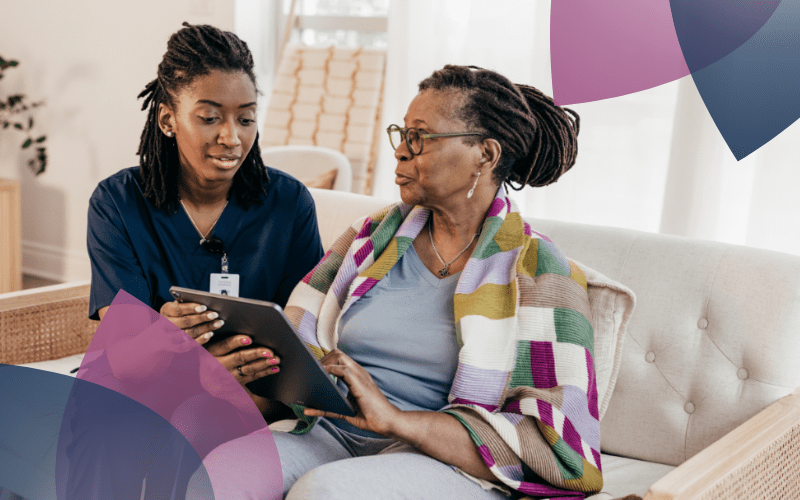 A support worker and an elderly participant sit together on a cosy couch in a well-lit home. The support worker, dressed in professional attire, attentively explains something on a tablet while the participant listens and engages with interest.