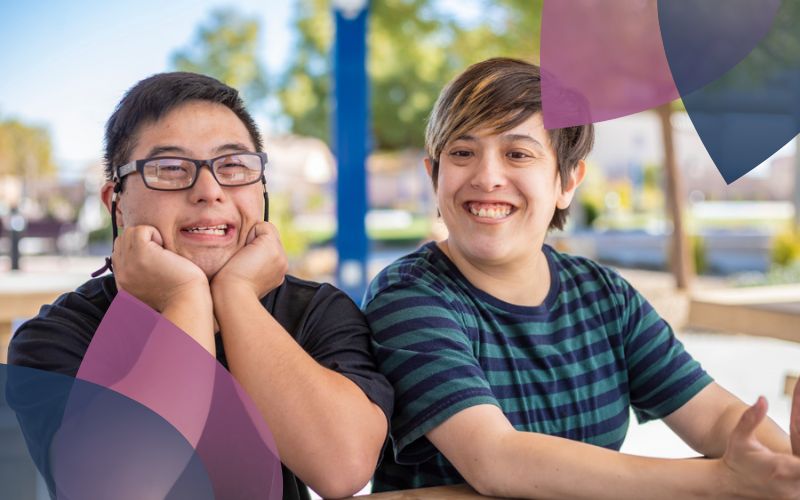 Two participants seated outdoors share a joyful moment, smiling brightly. The person on the left rests their hands on their cheeks, while the person on the right leans slightly towards them. The background features a sunny park-like setting with trees and pathways, creating a relaxed and inclusive atmosphere.