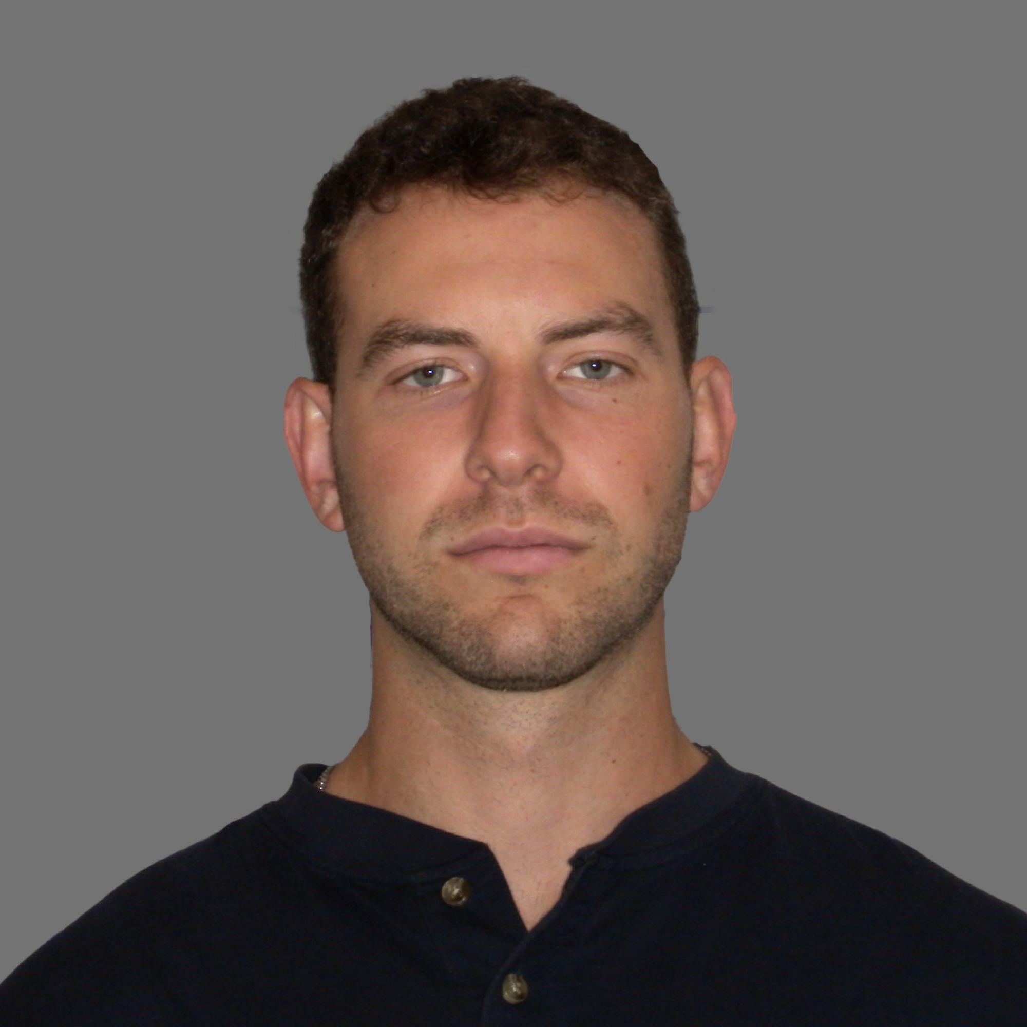 Portrait of a young man with short curly brown hair, light stubble, wearing a dark buttoned shirt against a gray background.