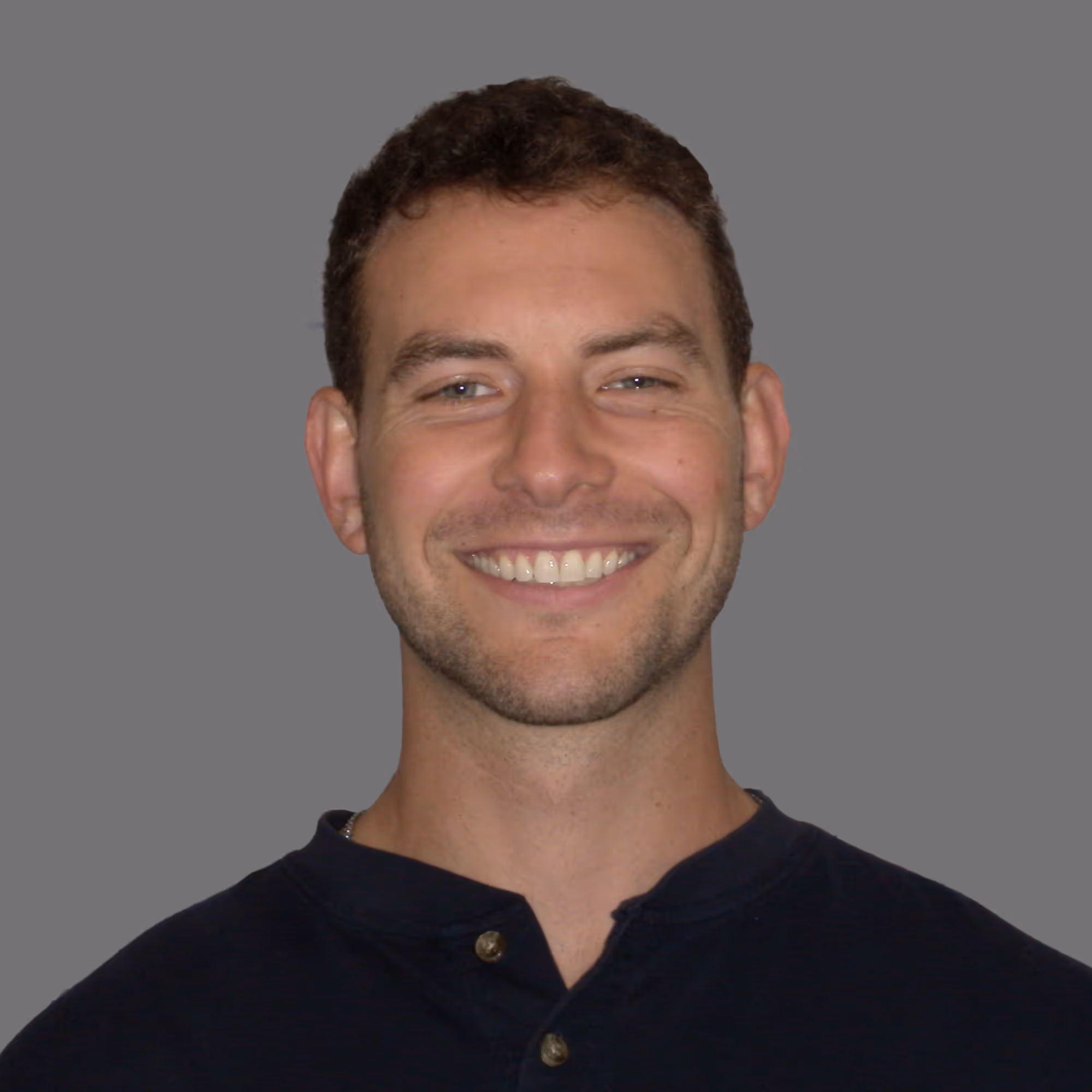 Smiling man with short curly hair wearing a dark buttoned shirt against a gray background.