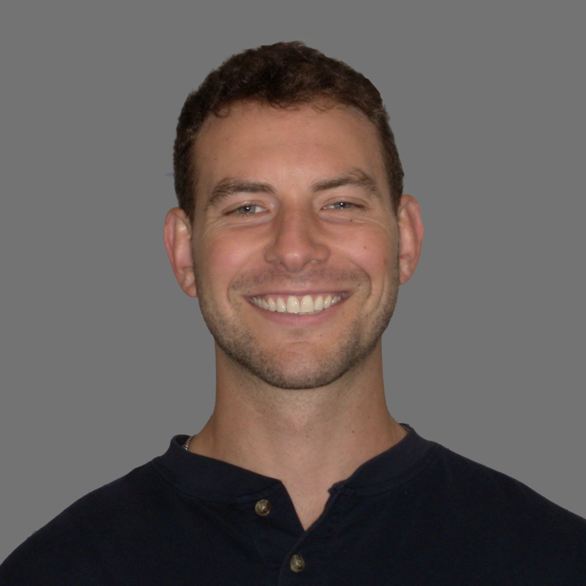 Smiling man with short curly hair wearing a dark buttoned shirt against a gray background.