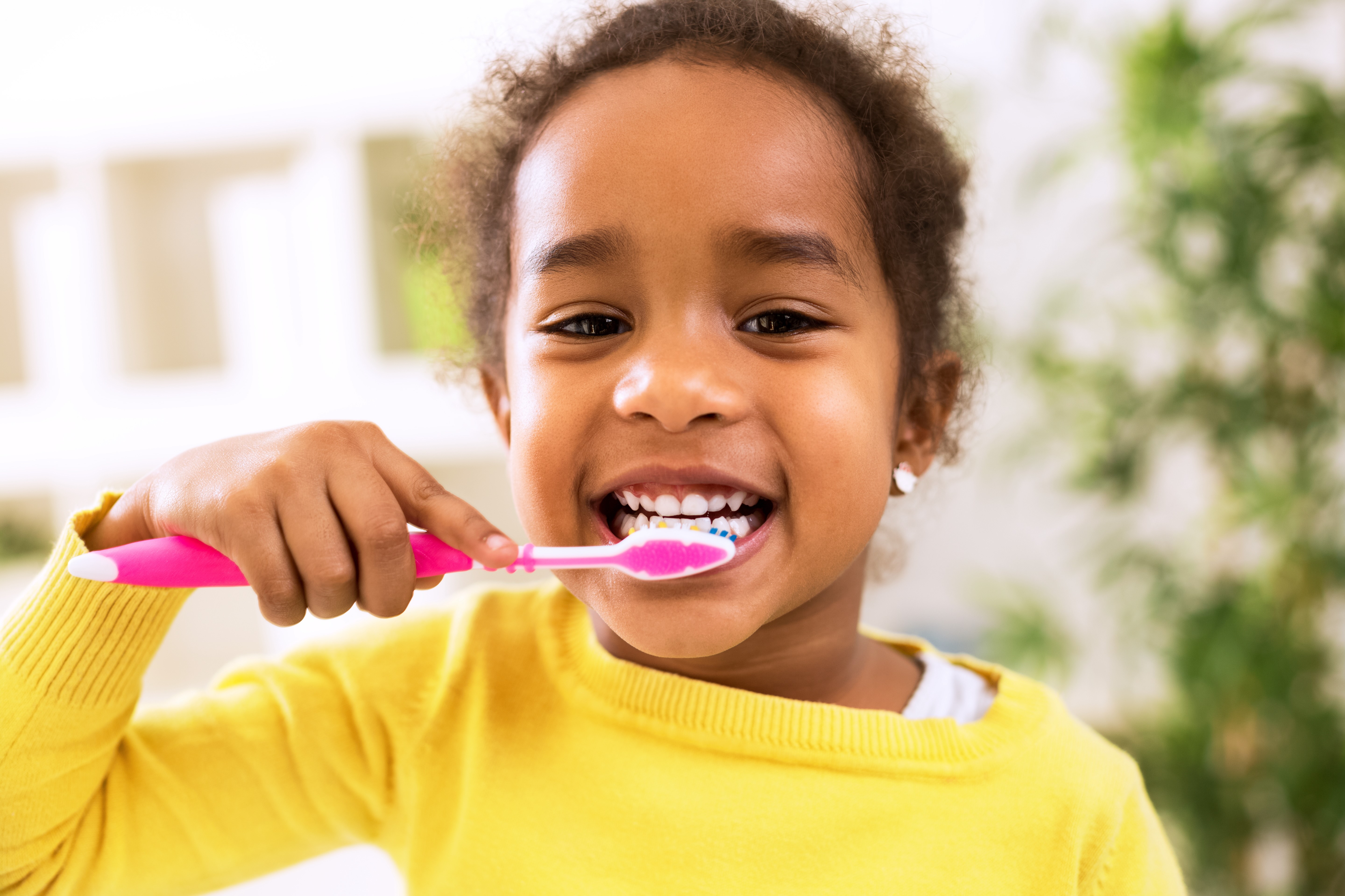 Young child smiling while brushing their teeth with a pink toothbrush.