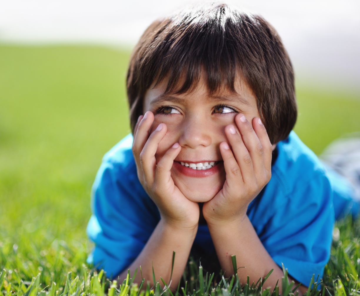Smiling young boy lying on green grass with hands resting on his cheeks, looking to the side.