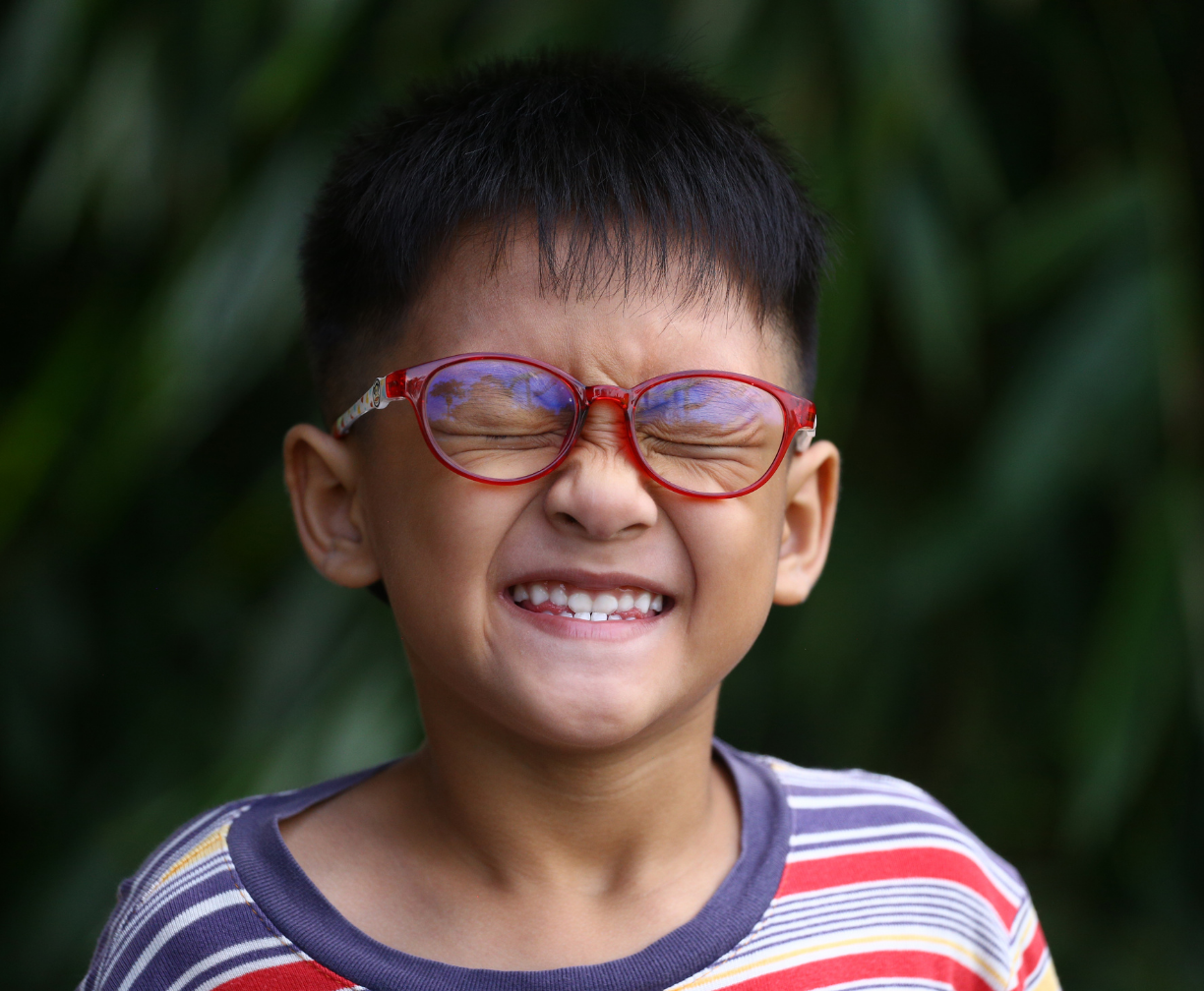 Young boy wearing red glasses and a striped shirt with eyes tightly closed and smiling.