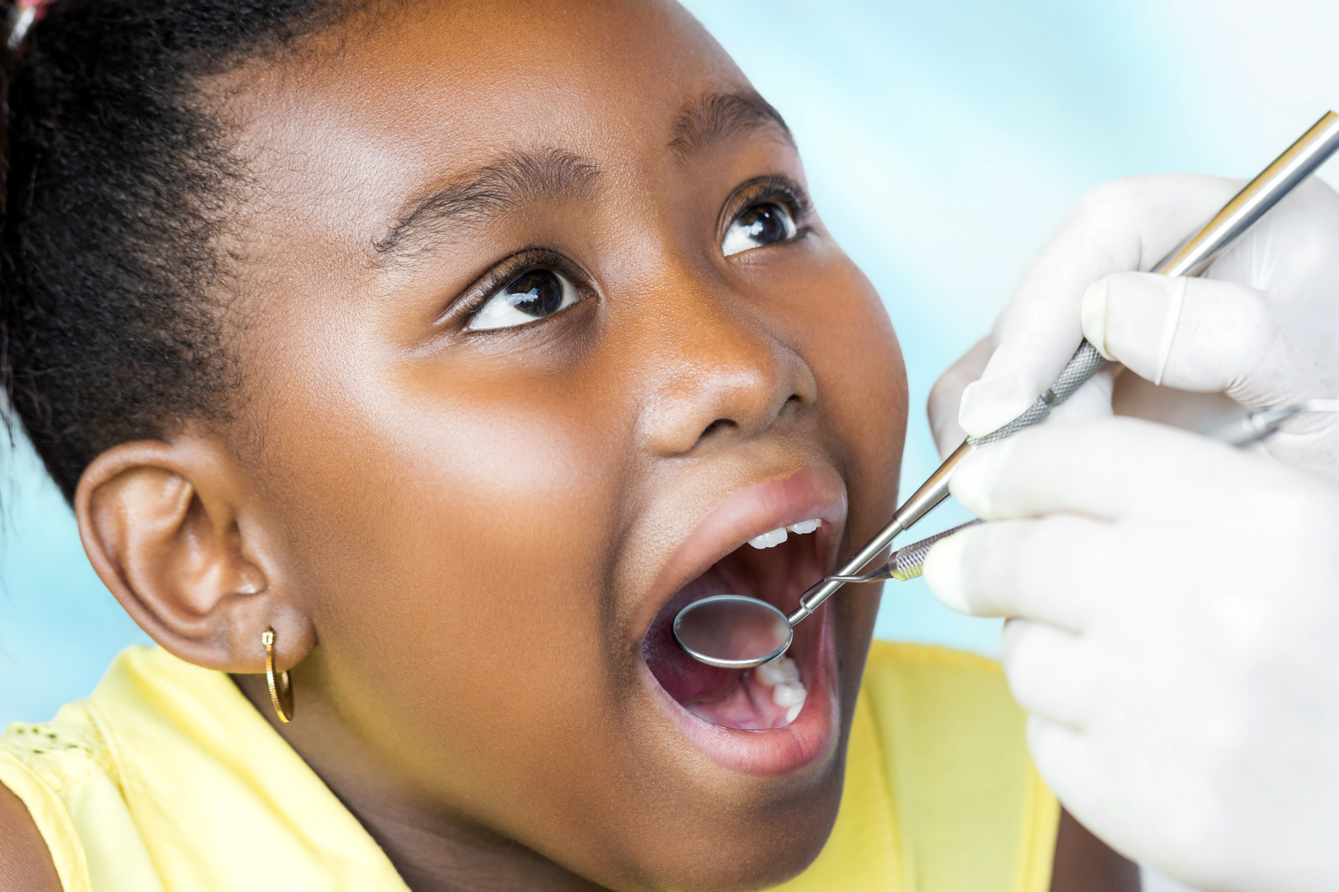 Girl with open mouth undergoing dental examination with dental mirror and explorer held by gloved hands.
