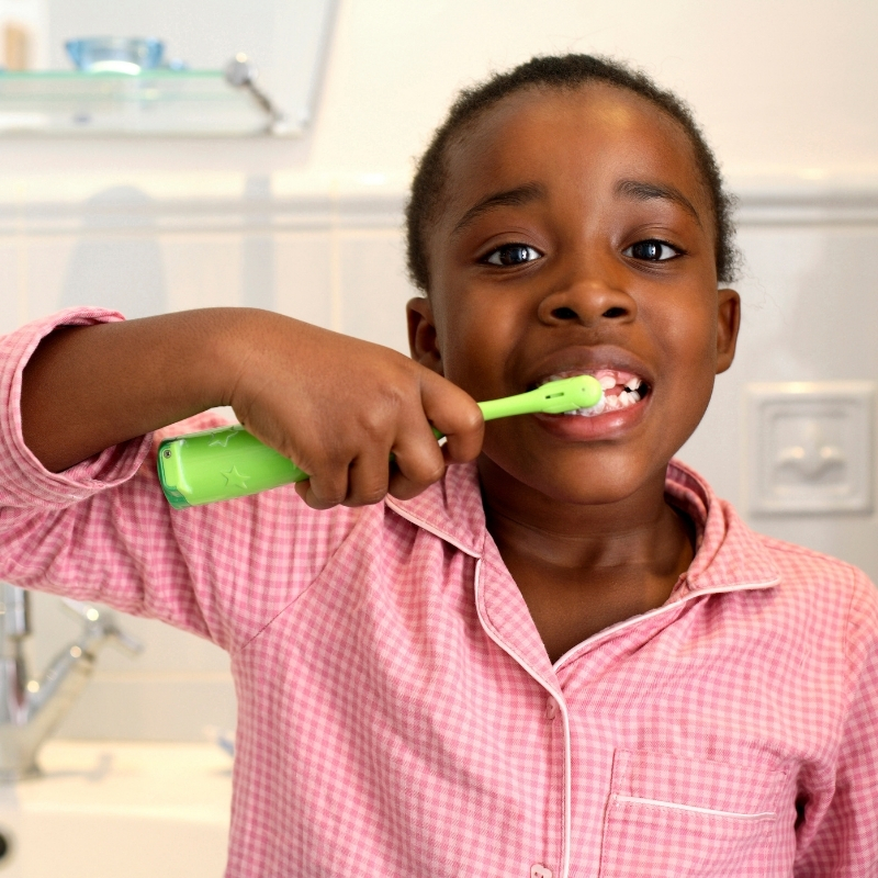 Smiling child in pink pajamas brushing teeth with a green toothbrush in a bathroom.
