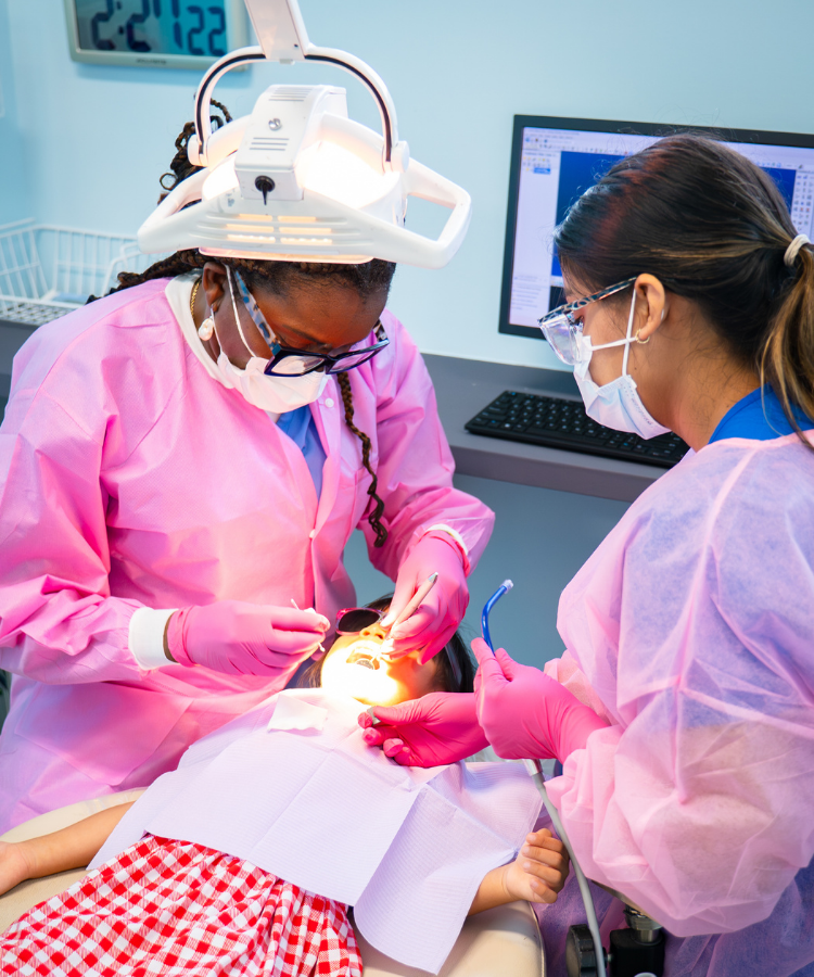 Two dental hygienists wearing masks and pink gowns examining a child in a red and white checkered dress in a dental chair.