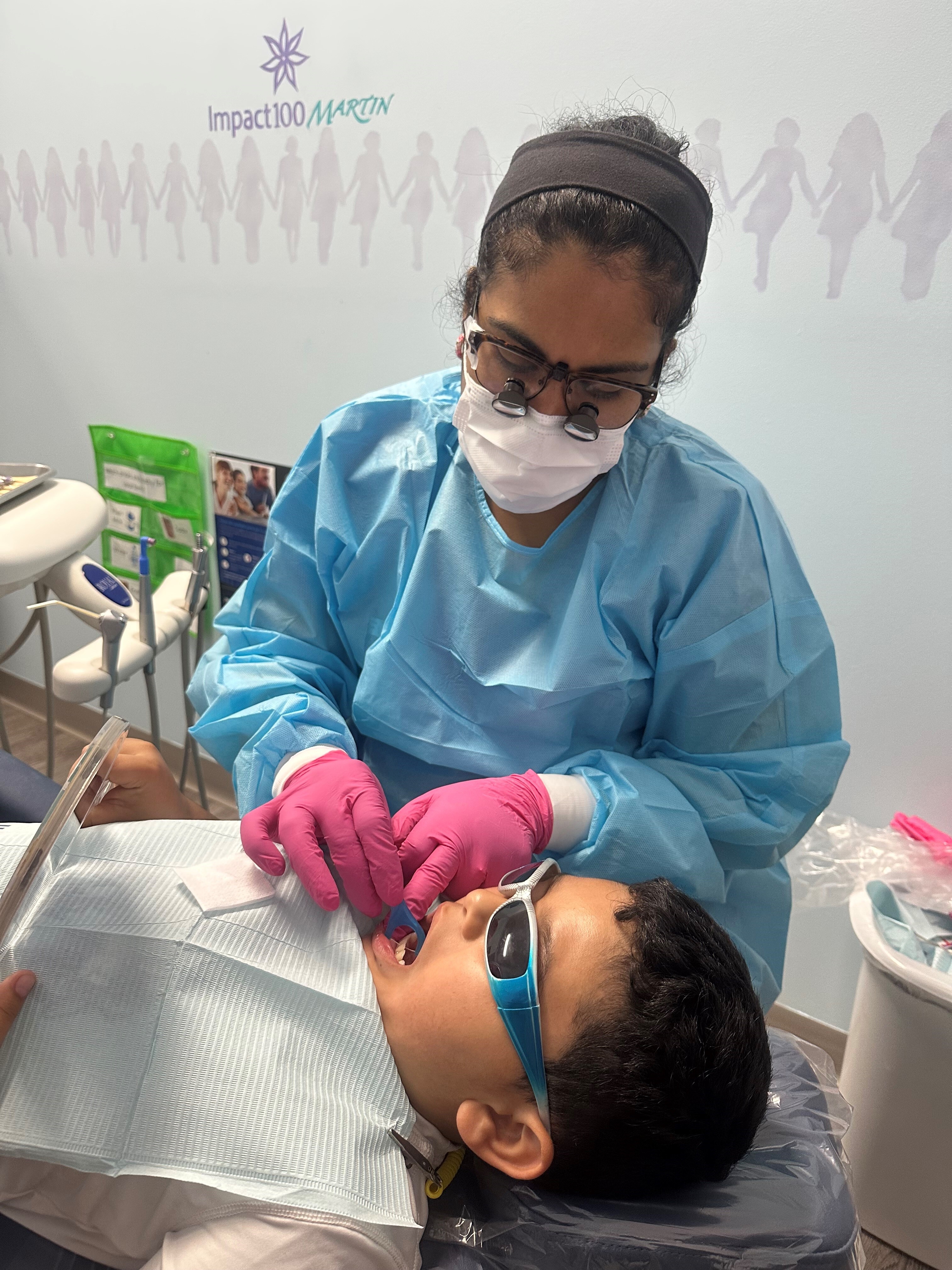 Dentist wearing magnifying glasses and pink gloves examining a young boy's mouth at a dental clinic.