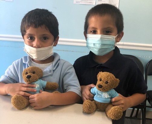 Two young boys wearing face masks, sitting at a table and holding teddy bears dressed in blue shirts.