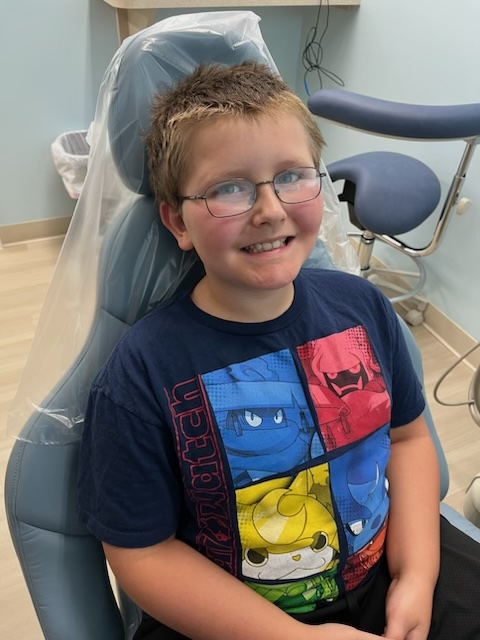 Smiling boy wearing glasses and a colorful cartoon ninja t-shirt sitting in a dental chair.