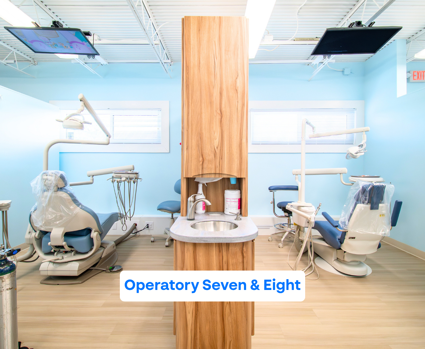 Bright dental operatory room with two dental chairs facing opposite directions and a wooden cabinet with a sink in the center.