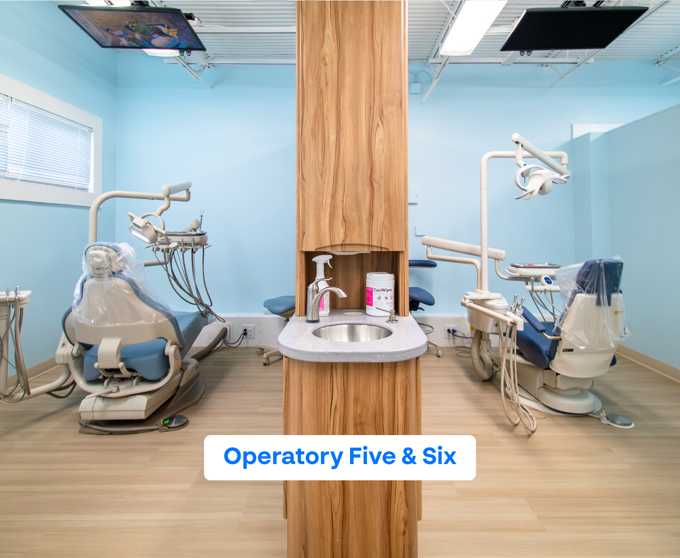 Two dental operatories separated by a wooden cabinet with a sink, each operatory featuring a dental chair and equipment against light blue walls.