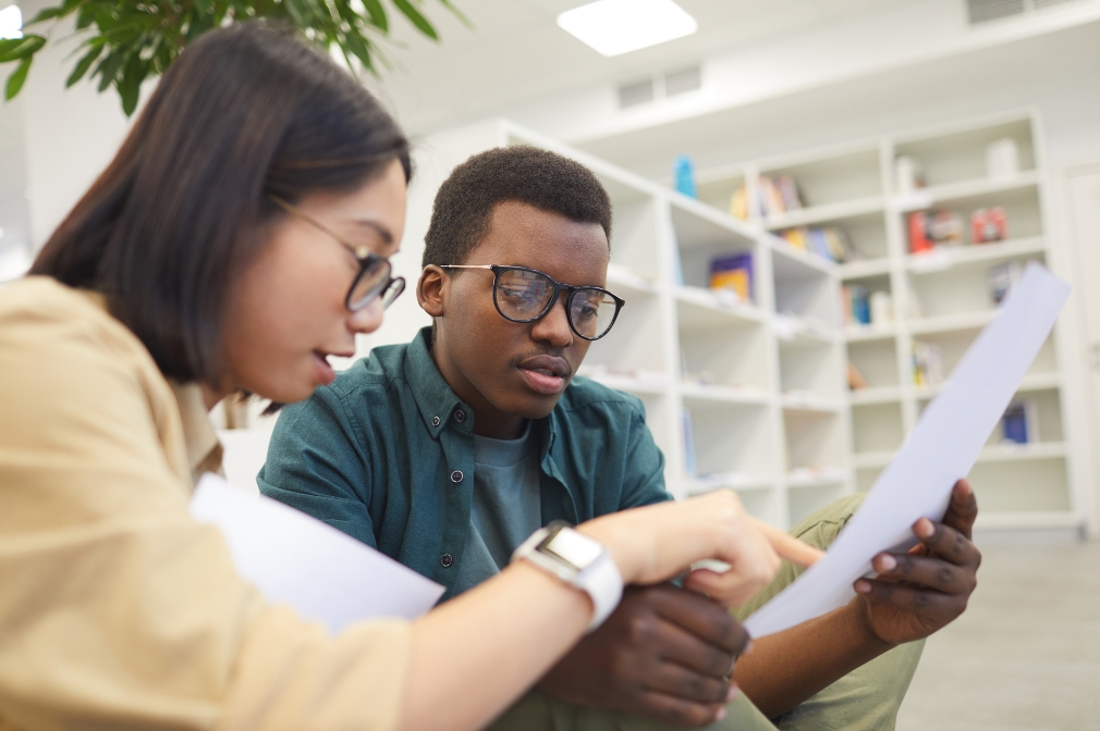 Two young people wearing glasses sitting indoors reviewing a document together.