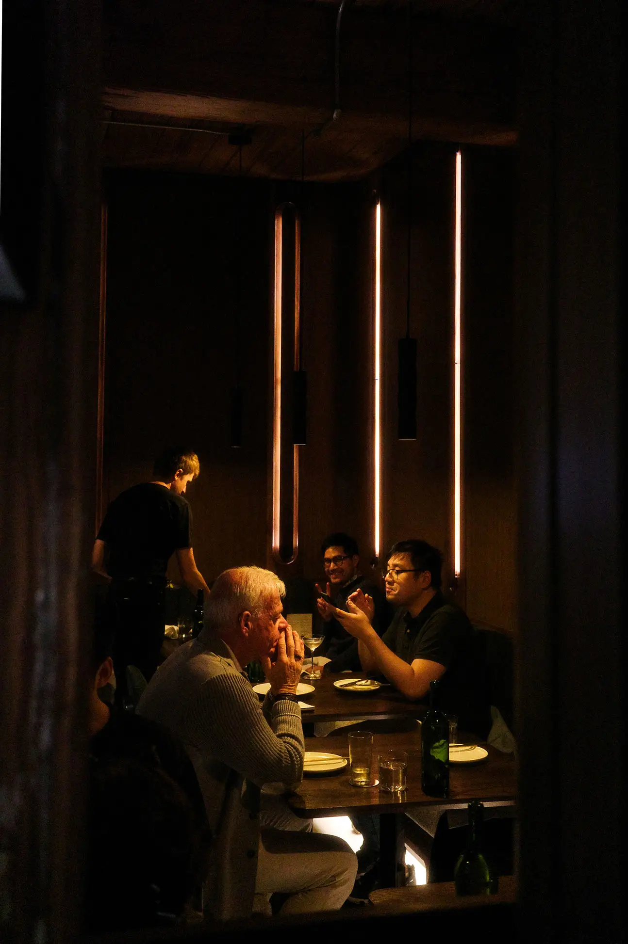 People sitting and conversing at a dimly lit restaurant table with vertical strip lights on the wall.