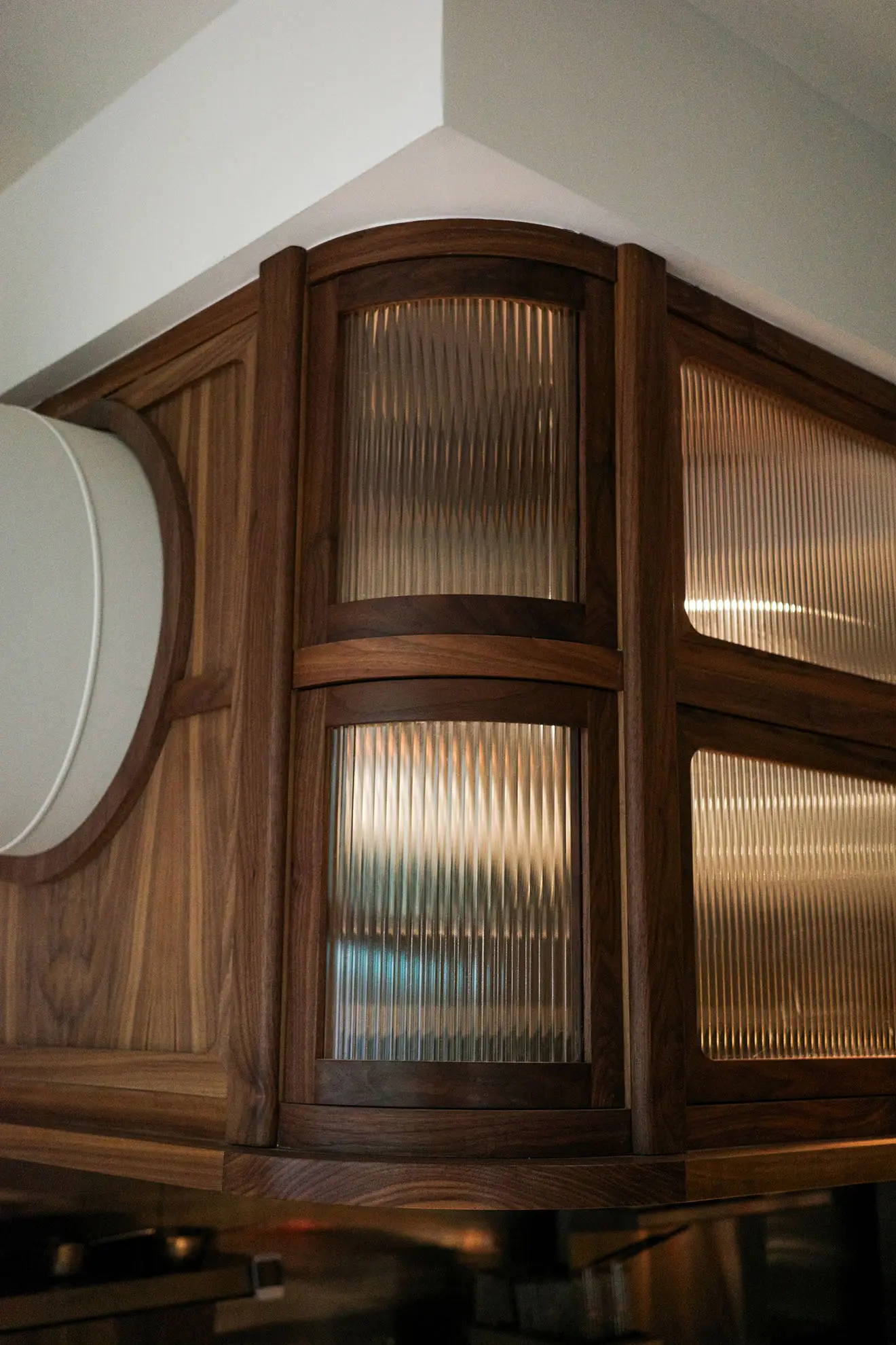 Close-up of a wooden corner cabinet with ribbed glass panels in warm ambient light.