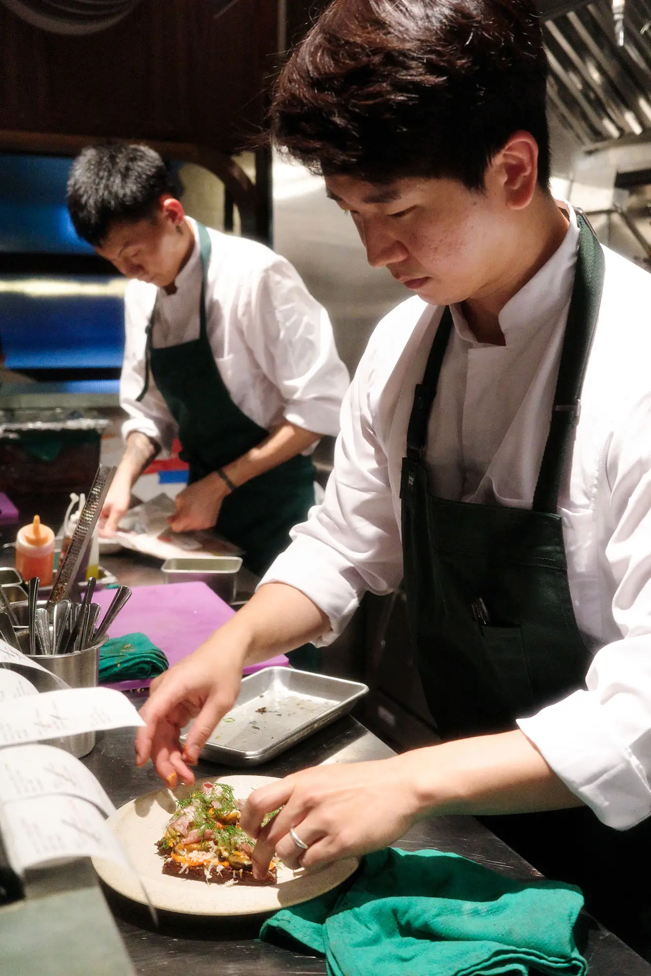 Chef in a white shirt and dark apron carefully plating a dish in a busy restaurant kitchen.