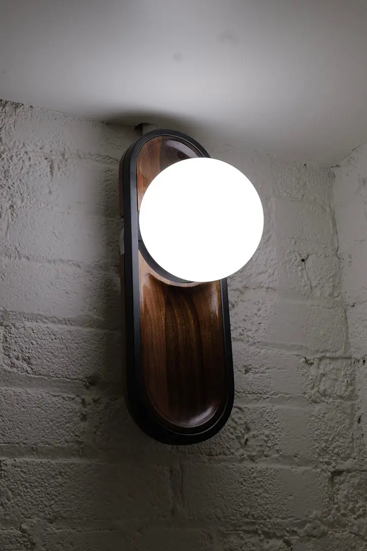 Modern wall-mounted light fixture with a round glowing bulb and wooden backplate on a textured white brick wall.