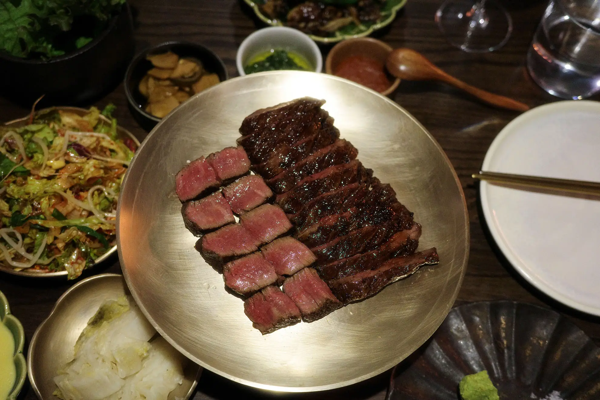Slices of medium-rare steak on a gold plate surrounded by small dishes of salad, sauces, and chopsticks on a wooden table.
