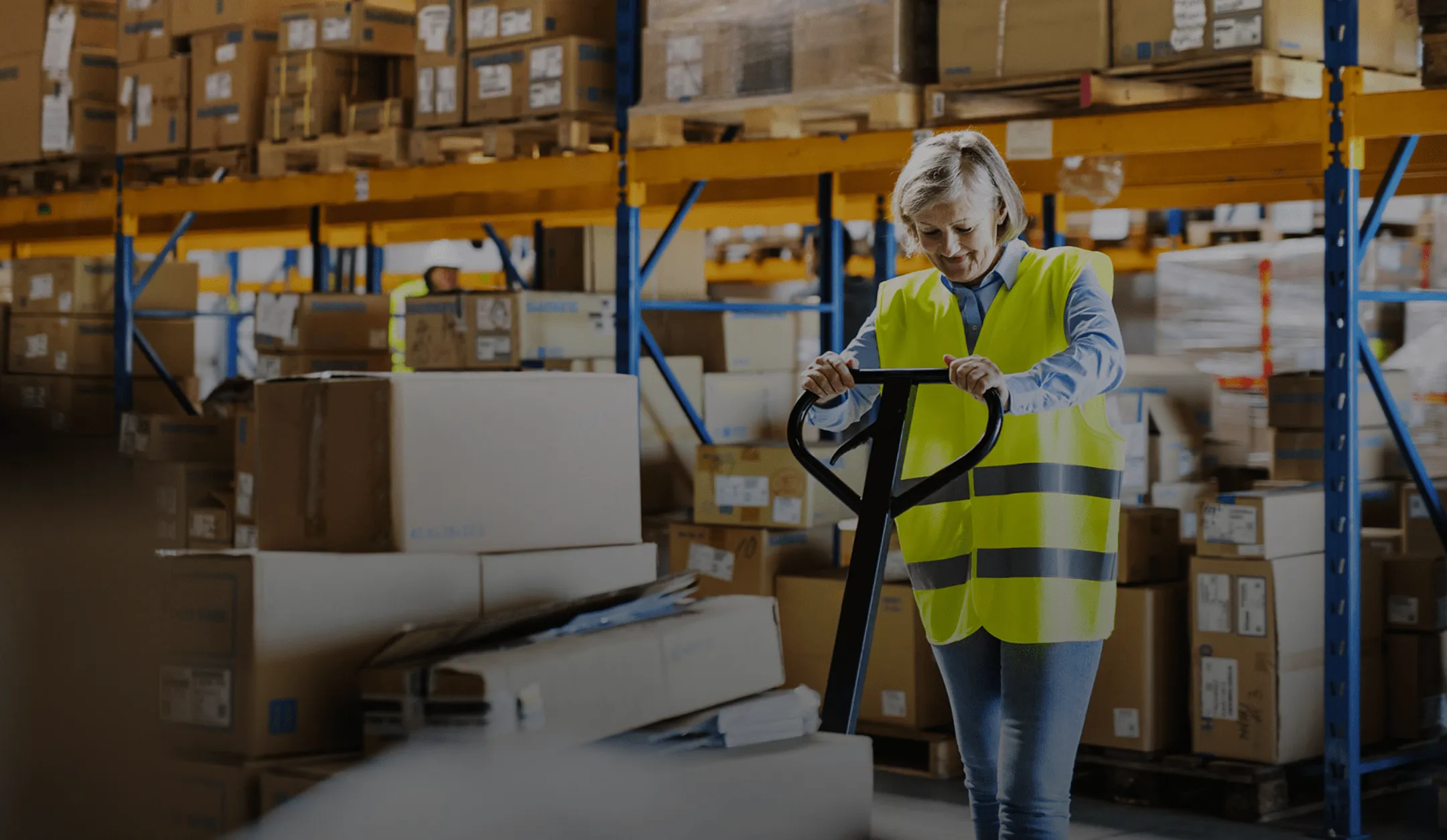 A woman in a yellow vest is pushing a cart in a warehouse.