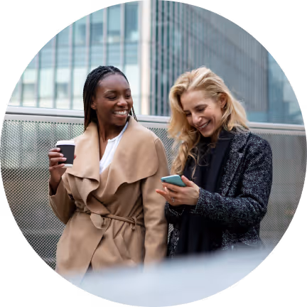 Two women wearing coats outside of an office building with one holding her mobile phone and another holding a cup of coffee.