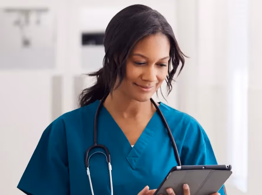Smiling female healthcare professional in teal scrubs using a tablet with a stethoscope around her neck.