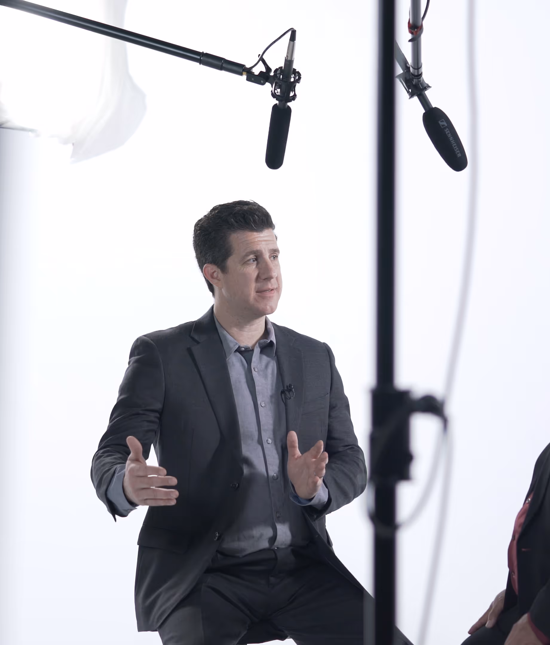 Man in a dark suit speaking and gesturing during an interview with two overhead microphones visible against a plain white background.
