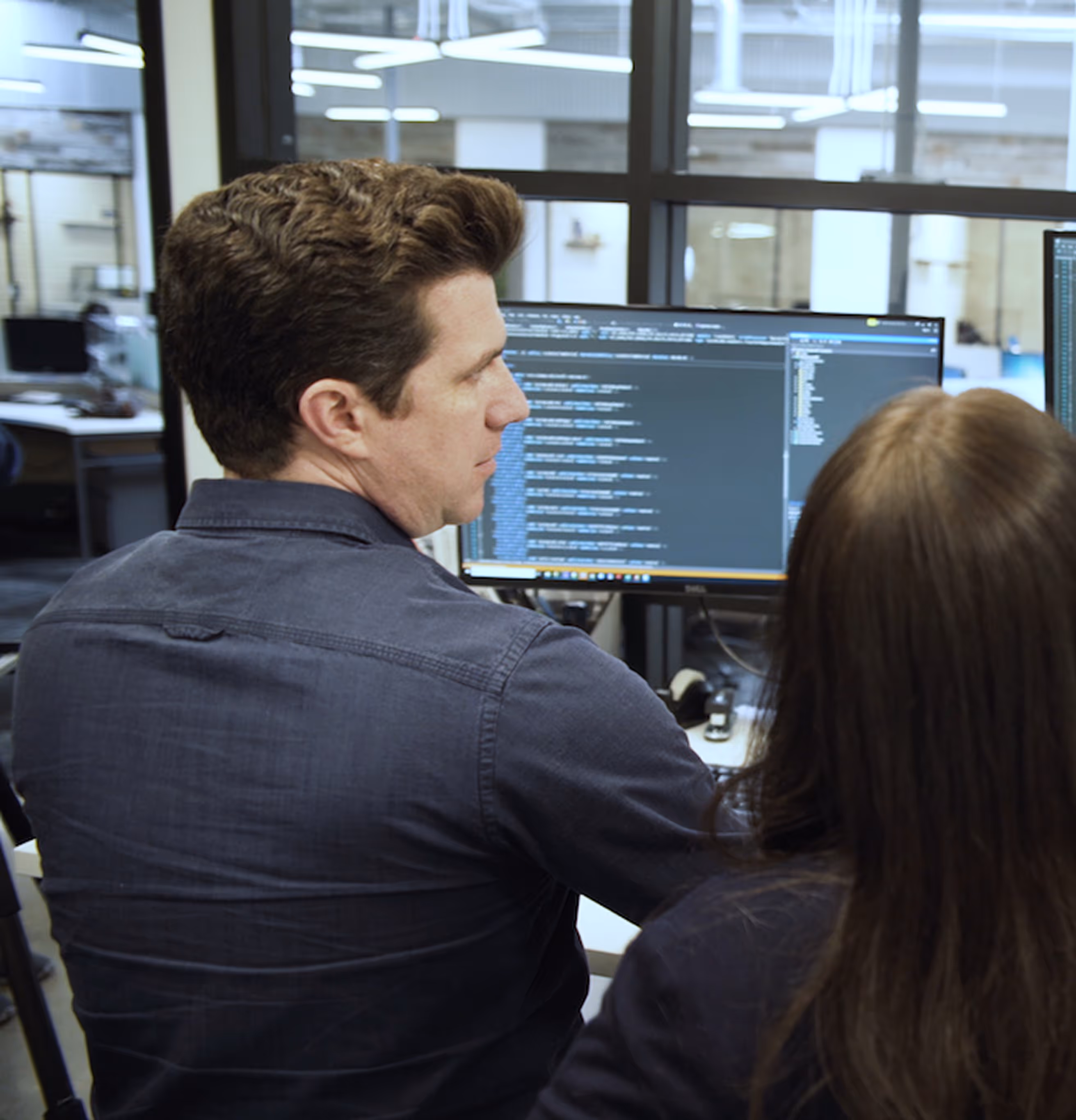 Two people working together at a desk in an office, looking at a computer screen displaying code.