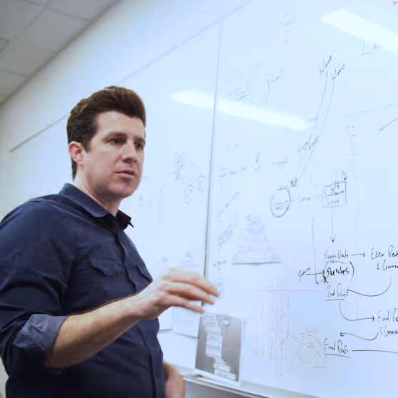 Man standing and gesturing in front of a whiteboard filled with diagrams and notes in an office setting.