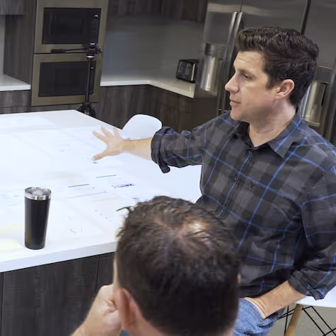 Man in a plaid shirt gesturing towards documents spread out on a white table while another person listens in a modern kitchen setting.
