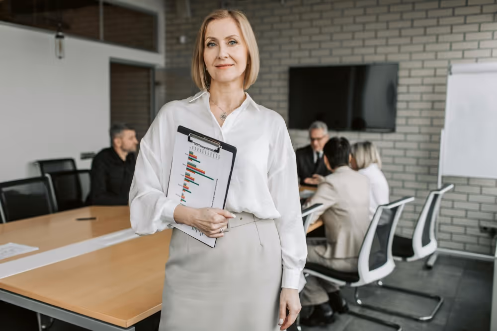 Confident woman in a white blouse holding a clipboard with a bar graph in a modern office, colleagues in discussion at a table behind her.