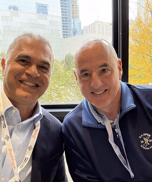Two men smiling indoors with window behind showing city buildings and autumn trees.