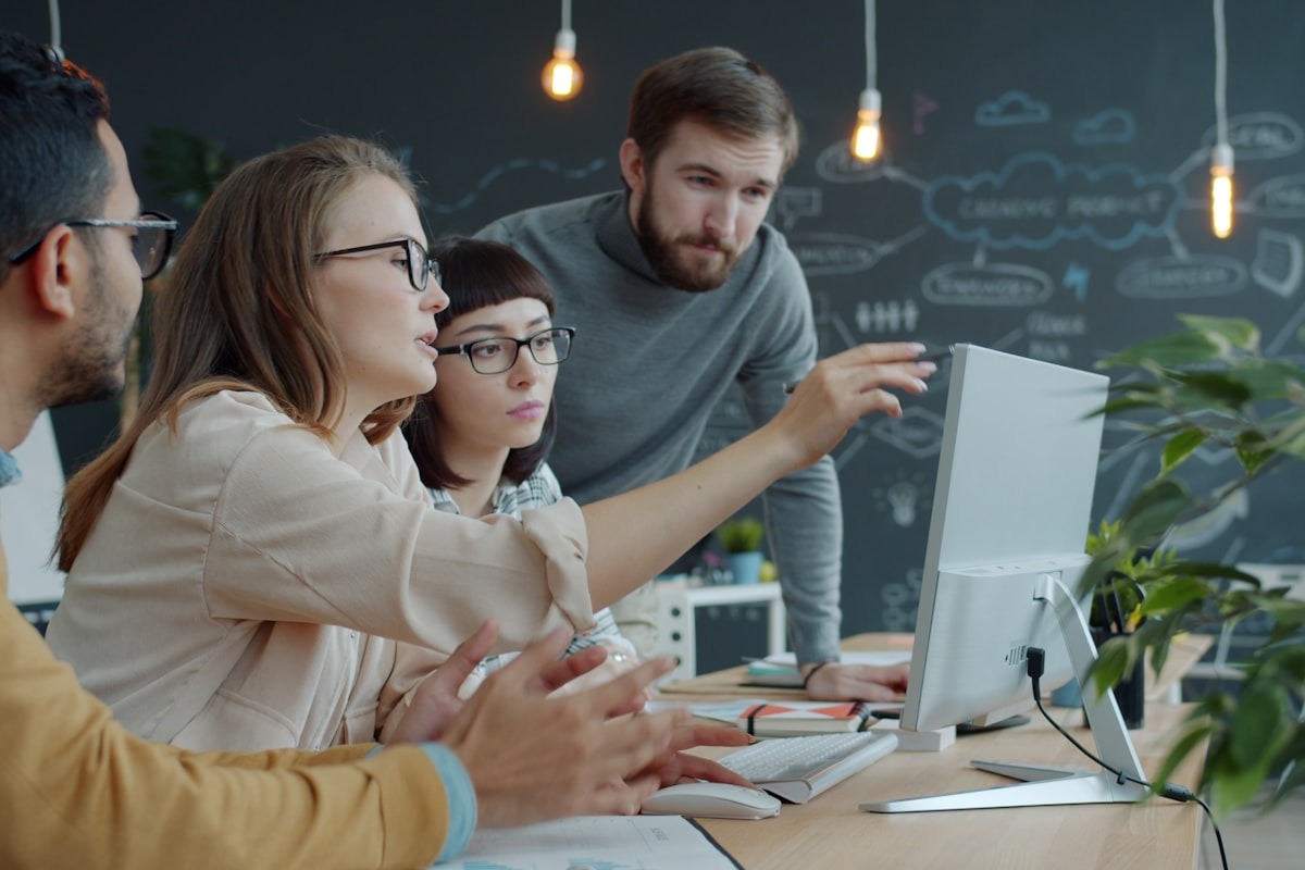 A diverse startup team of four people collaborating around a computer monitor in a creative office with a chalkboard wall