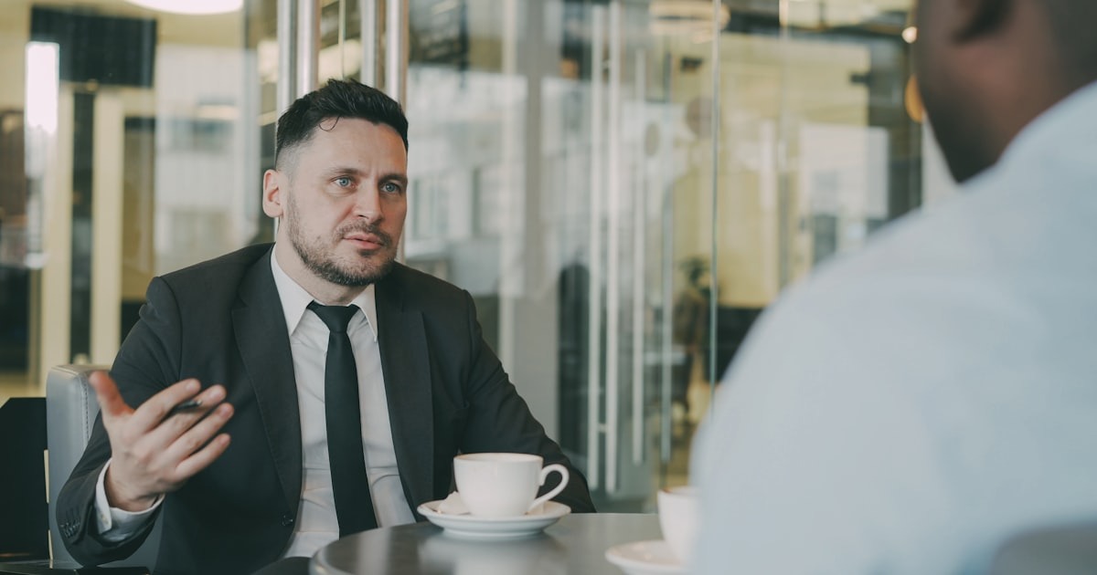 Two founders in a candid, exploratory conversation across a table, the kind of fit-finding call that precedes a coaching engagement.