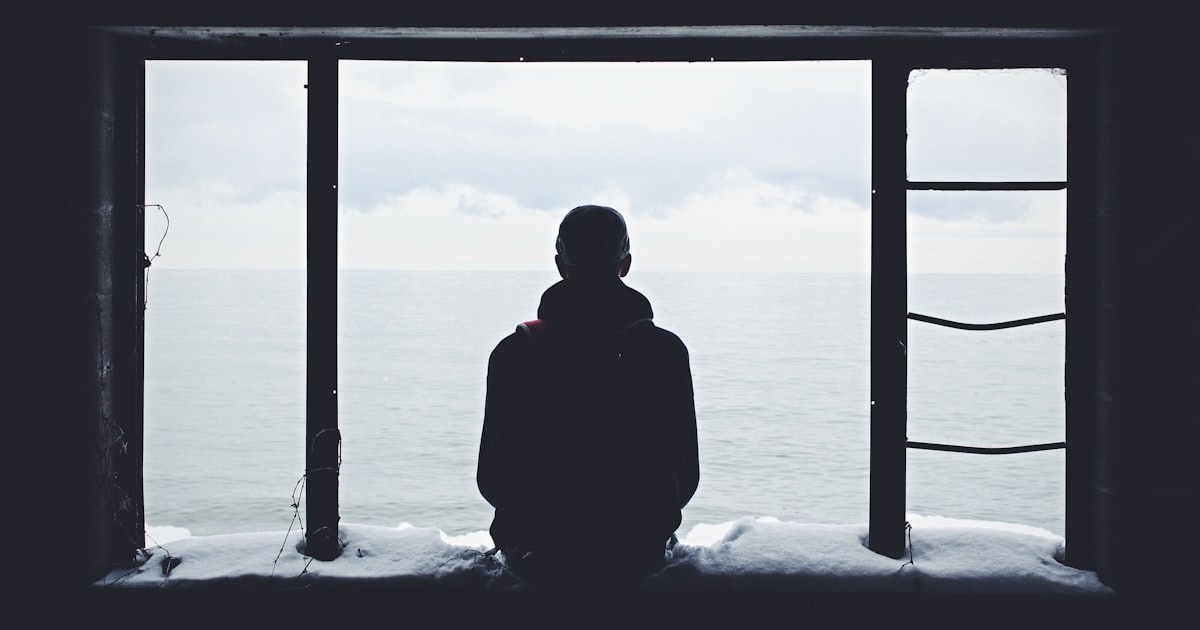 A lone figure sitting at a window looking out at the sea, evoking the isolation of making decisions without a peer.
