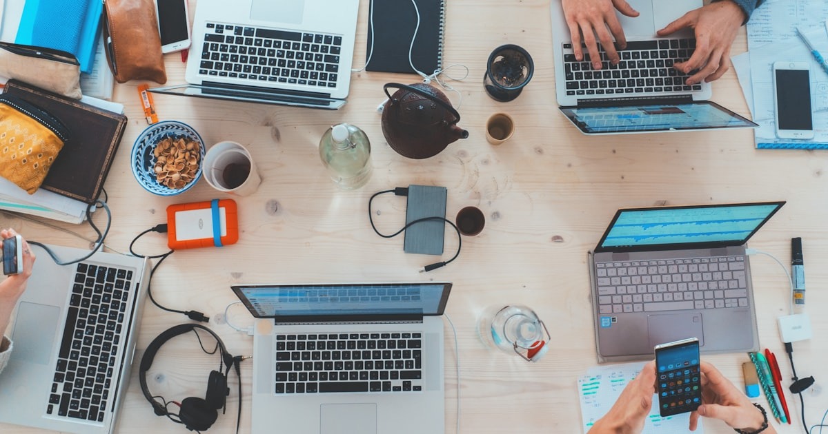 A shared desk crowded with open laptops, phones, cables, and coffee cups arriving from every direction, capturing the always-on chaos of reactive startup operating.