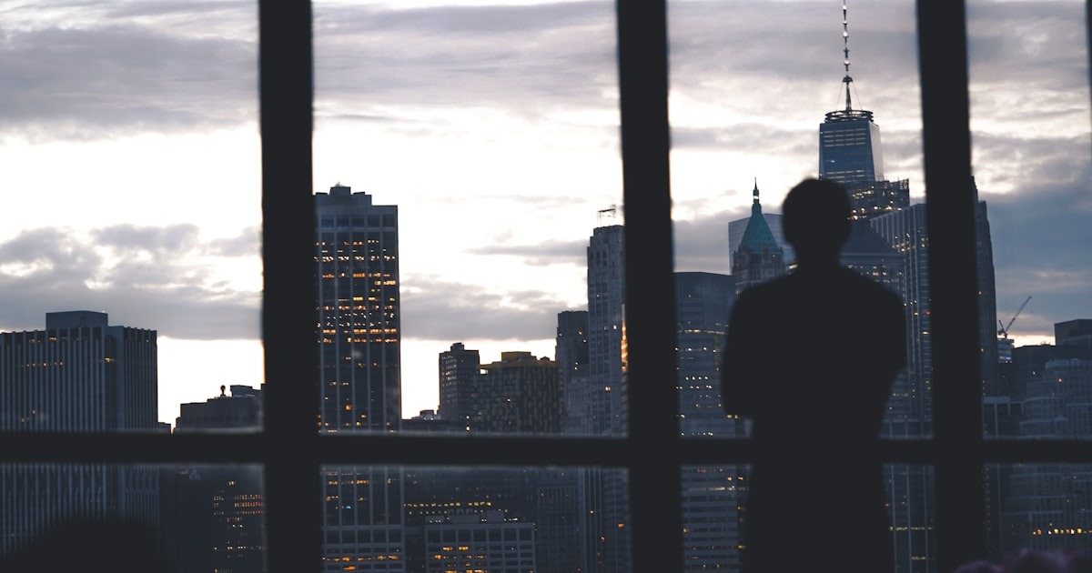 A founder standing calmly at a high-rise window over the city skyline, reflecting under pressure without cliché stress.