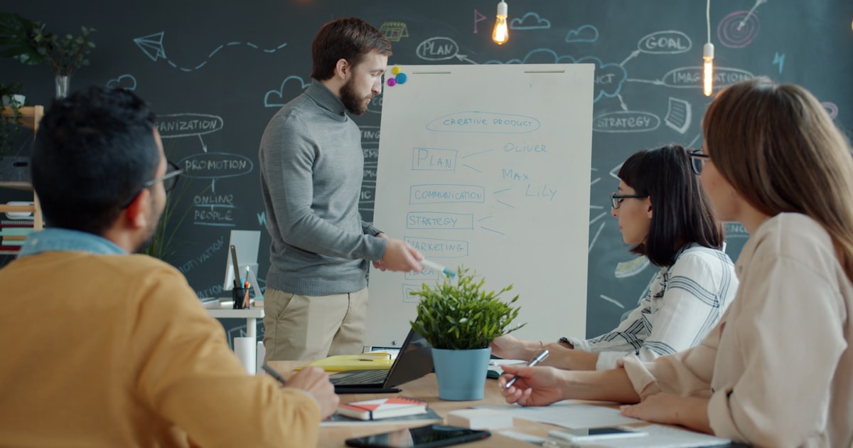 A team lead presents at a whiteboard while colleagues discuss strategy, illustrating a new coordination layer forming as a startup grows.