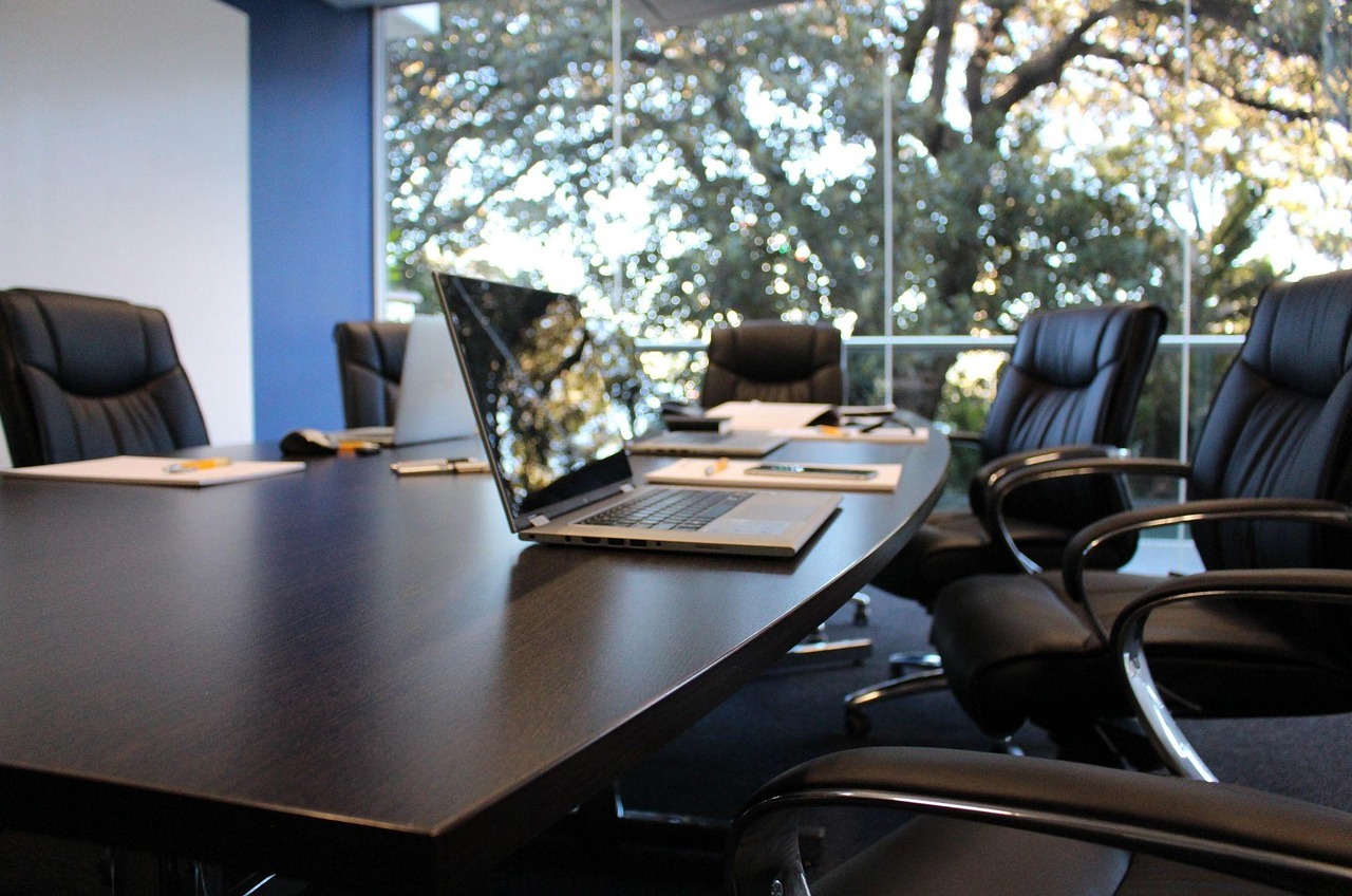 A modern glass-walled boardroom with a laptop open on a long table, chairs empty