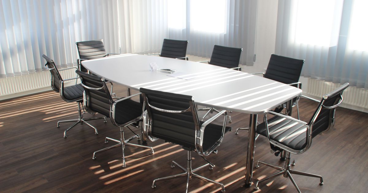 An empty meeting room with a white oval table, black chairs and low sunlight striped across a dark wood floor.
