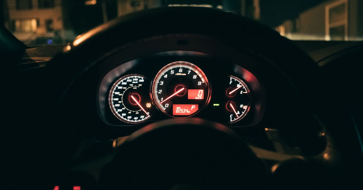 An illuminated car instrument cluster glows behind a steering wheel at night, with the speedometer reading zero.