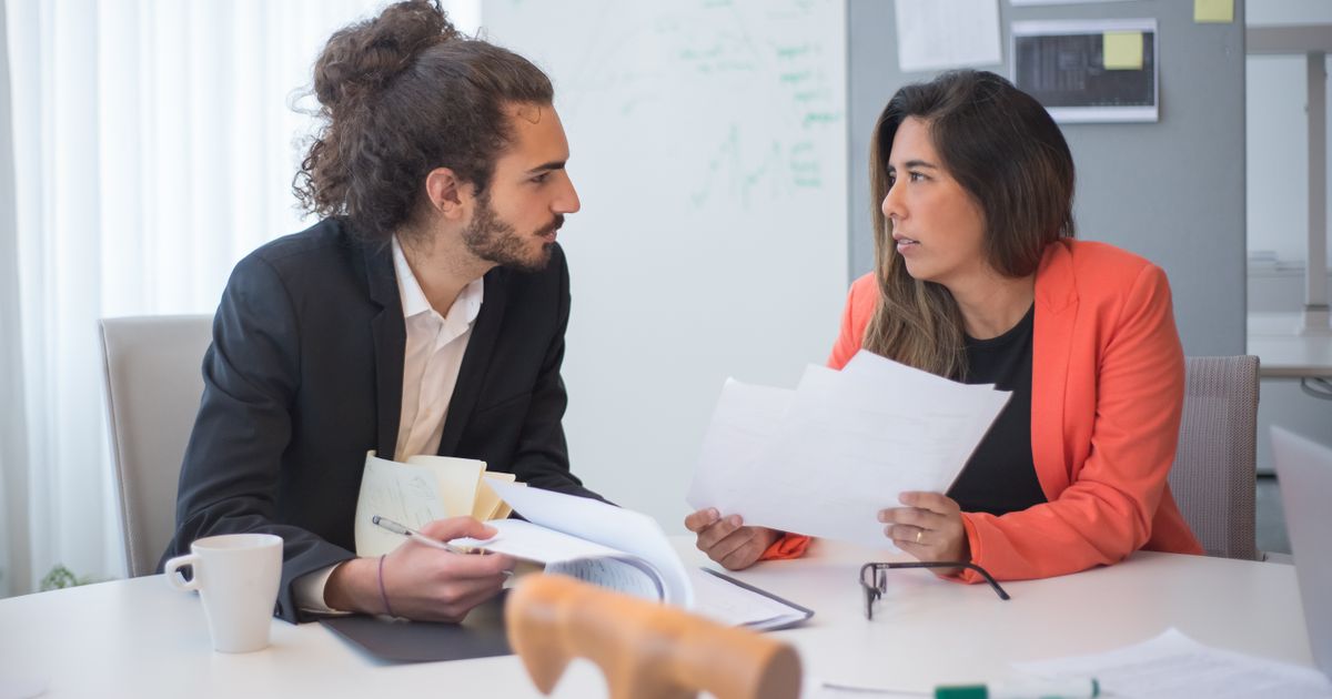 Two colleagues at a white table turn toward each other in conversation, one holding up printed pages.