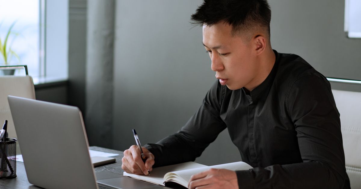 A man in a black shirt writes in an open notebook at a desk beside an open laptop, looking down at the page as he writes.