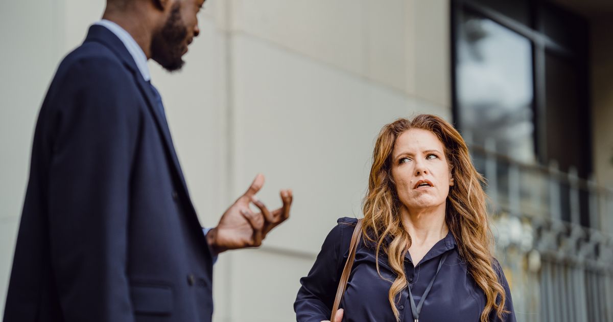 Two colleagues stand outside an office building, one gesturing mid-sentence while the other, wearing a lanyard, turns to look at him with a sceptical expression.
