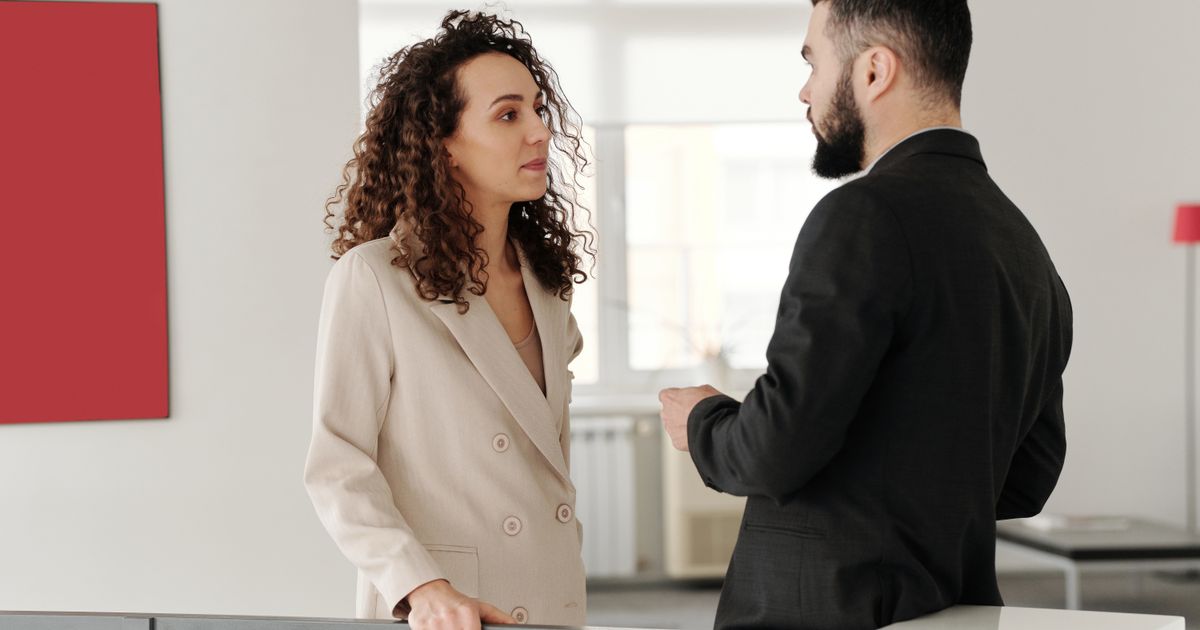 Two colleagues stand talking in a bright white office space, one listening with her hands resting on a railing while the other gestures mid-sentence.