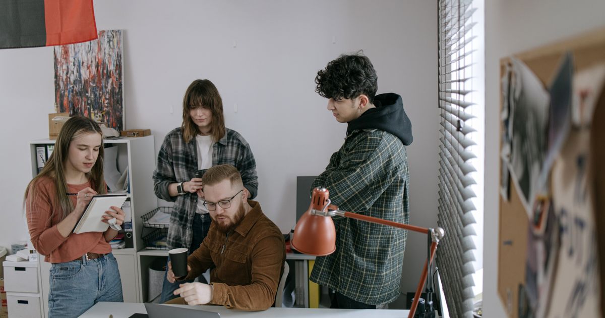 Four people gather around a desk in a small office with a bookshelf and an orange lamp, one seated at a laptop while a woman takes notes.